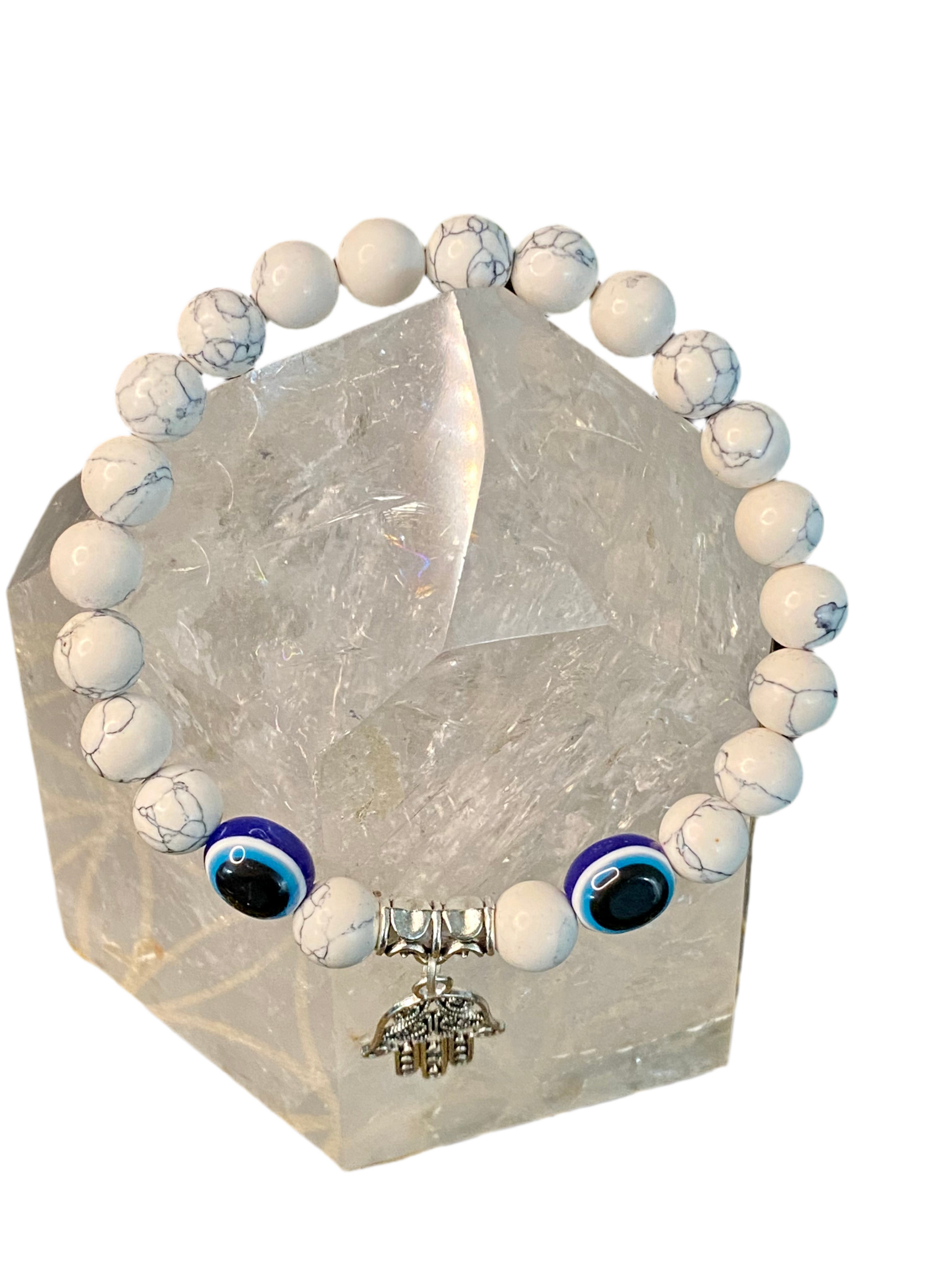 Howlite Evil Eye Gemstone Bracelet with Hamsa Charm on clear crystal stand.