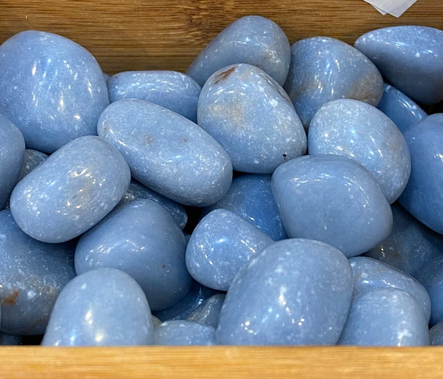 Angelite tumbled stones in a wooden box, highlighting smooth blue surface and high quality.